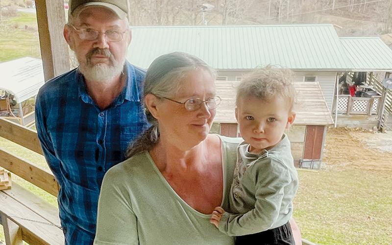Bobby and Rachel Riddle and their 22-month-old granddaughter Kenley are on the porch of their  daughter’s home, with their fire-damaged home in the background.
